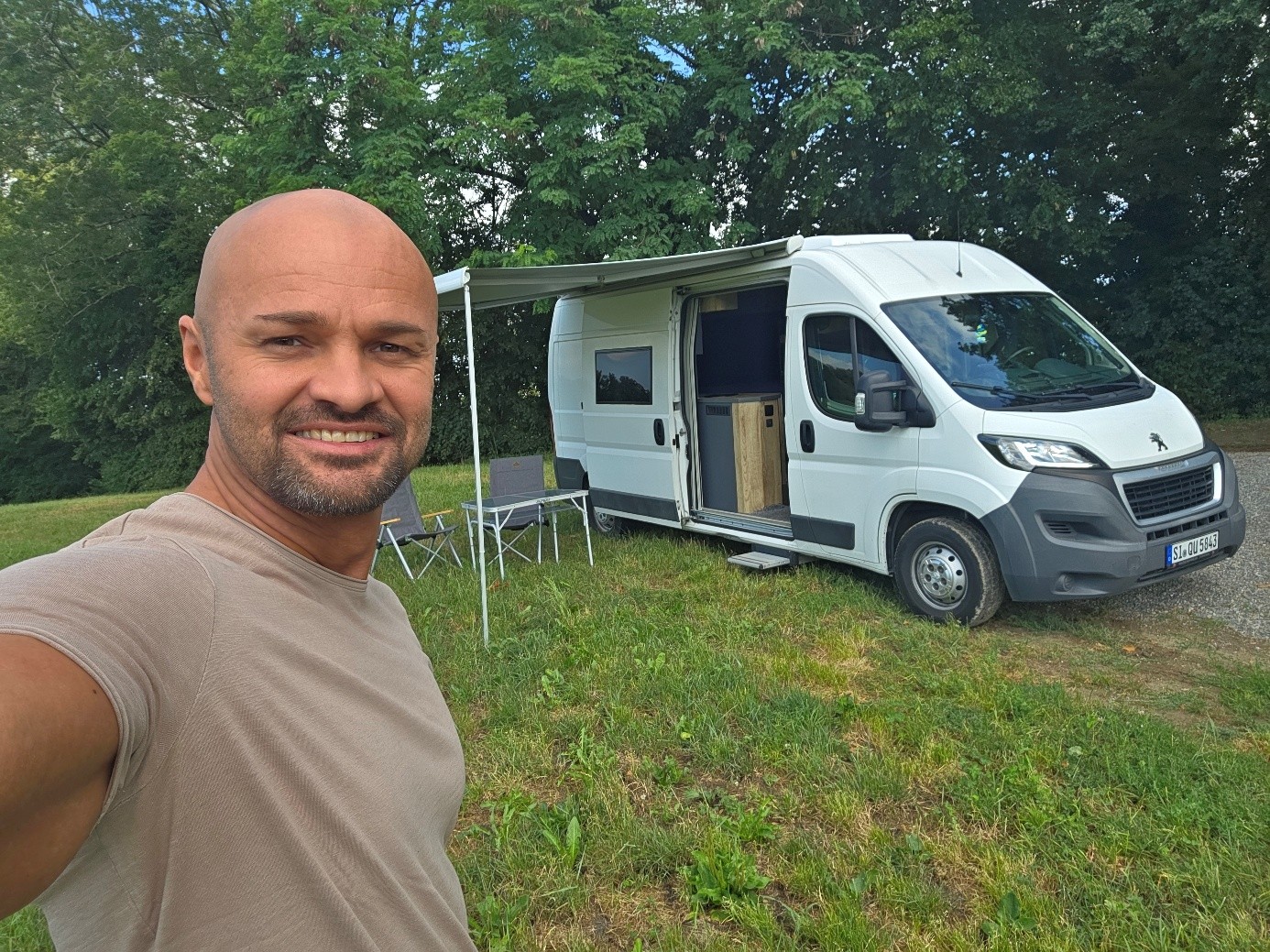 Eduard macht ein Selfie vor seinem Camper mit Markise auf einer grünen Wiese.