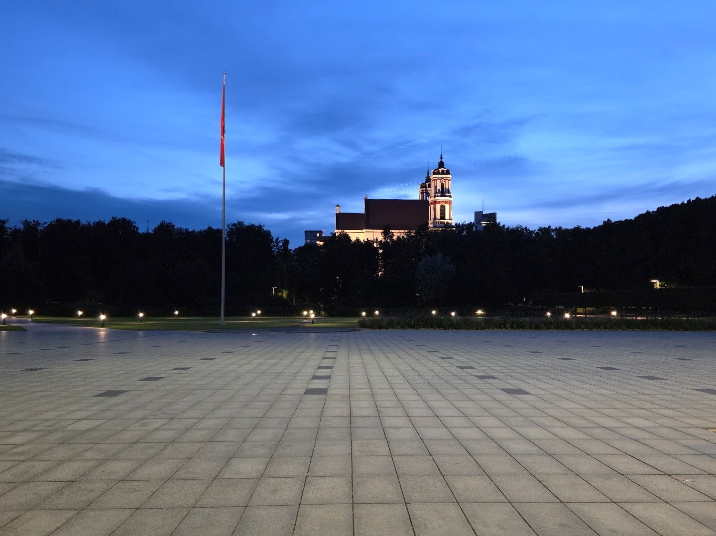 Gepflasterter Platz in der Dämmerung mit Fahnenmast und beleuchteter Kirche im Hintergrund.