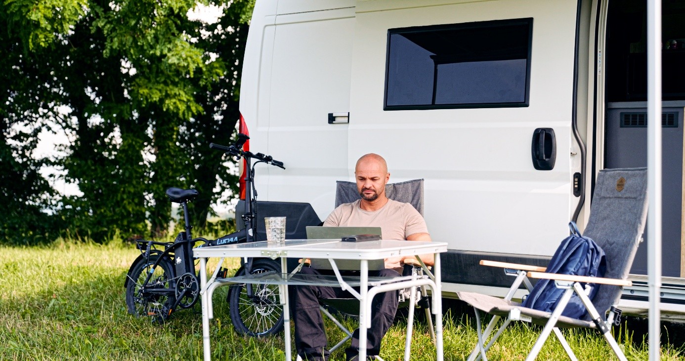 Eduard arbeitet am Laptop an einem Campingtisch vor seinem Camper im Grünen.
