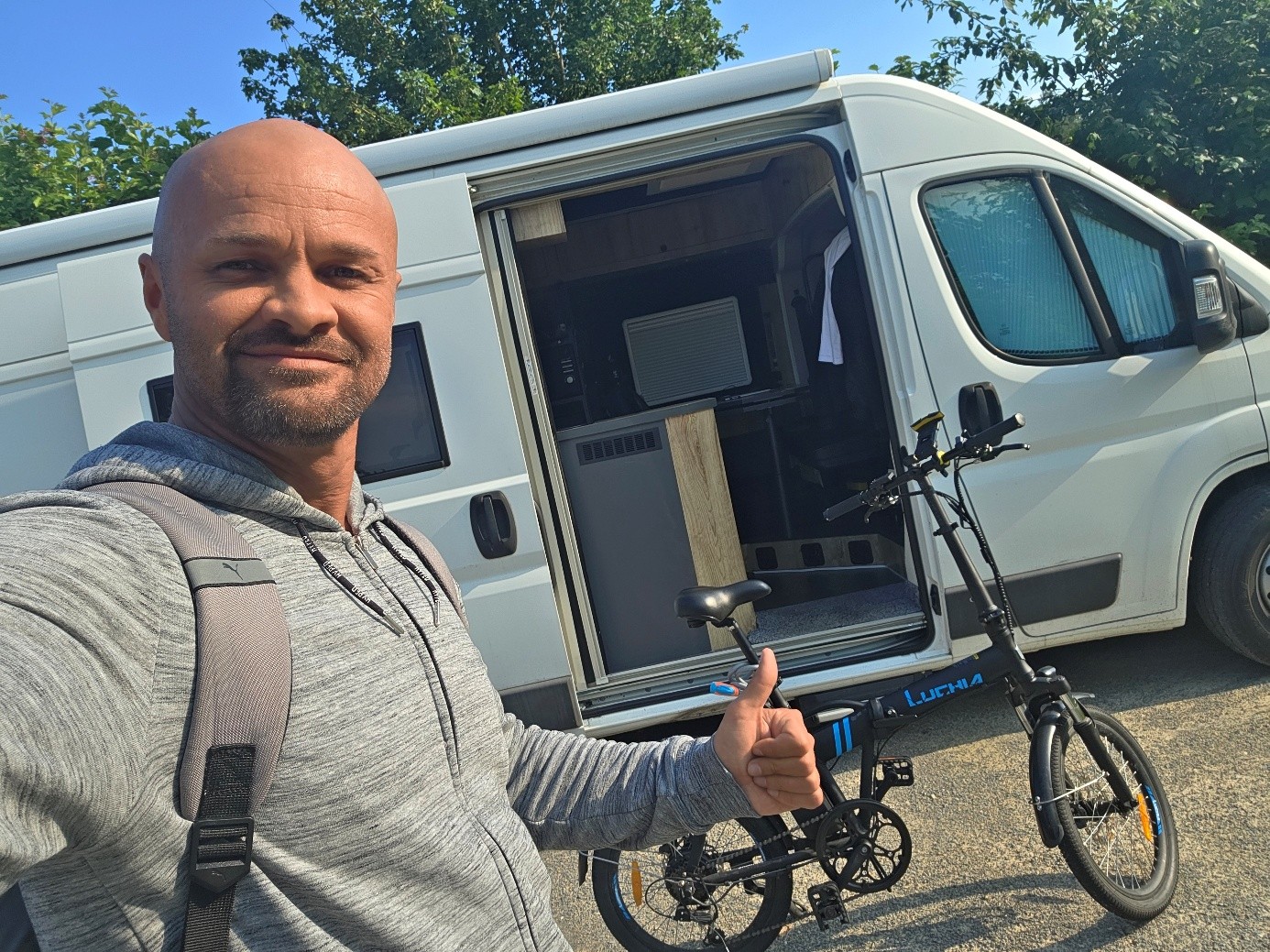 Eduard steht mit Daumen hoch neben einem schwarzen Faltrad vor seinem weißen Camper.