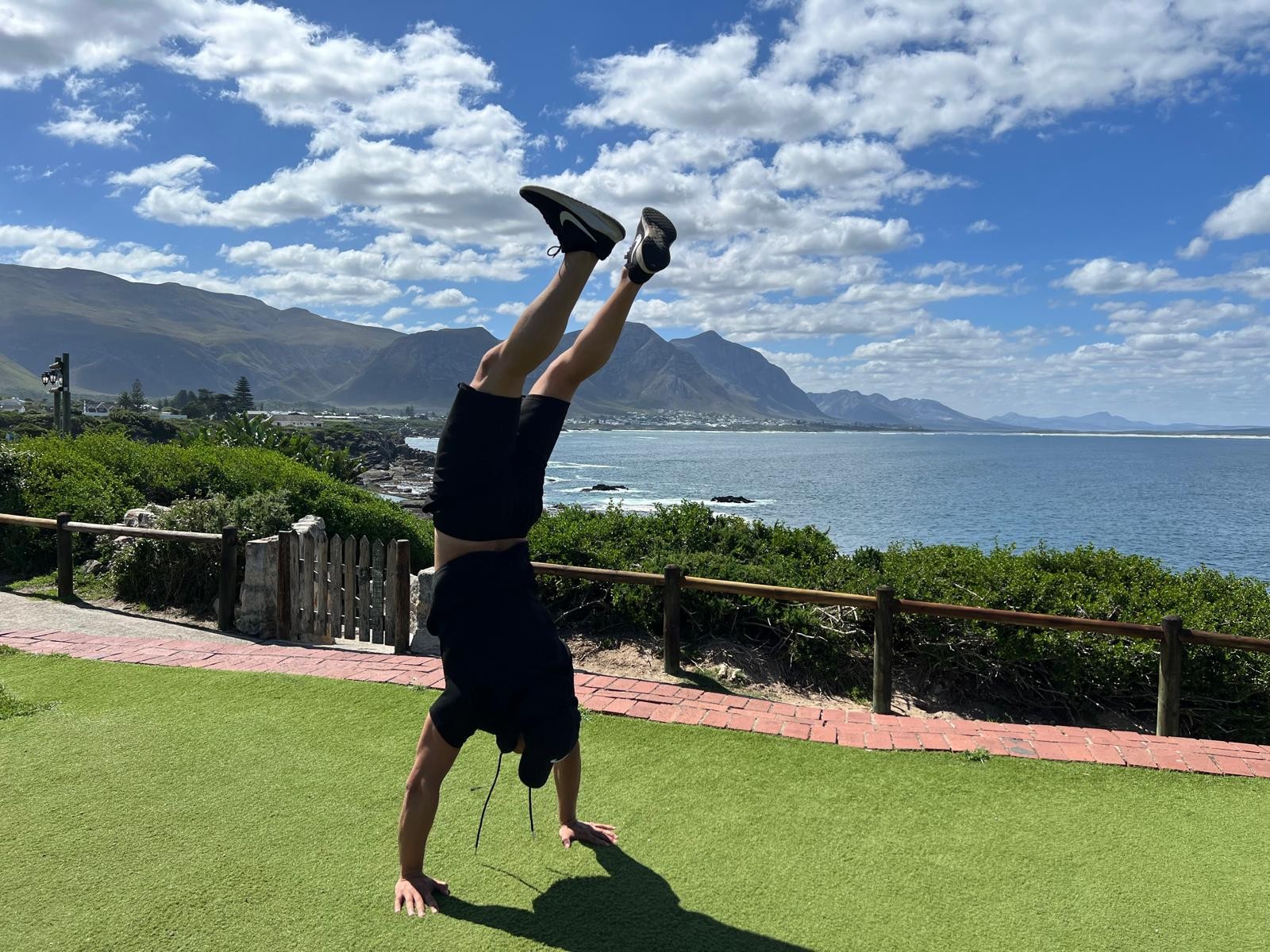 Eine Person macht einen Handstand auf einer Rasenfläche mit Blick auf Ozean und Berge.