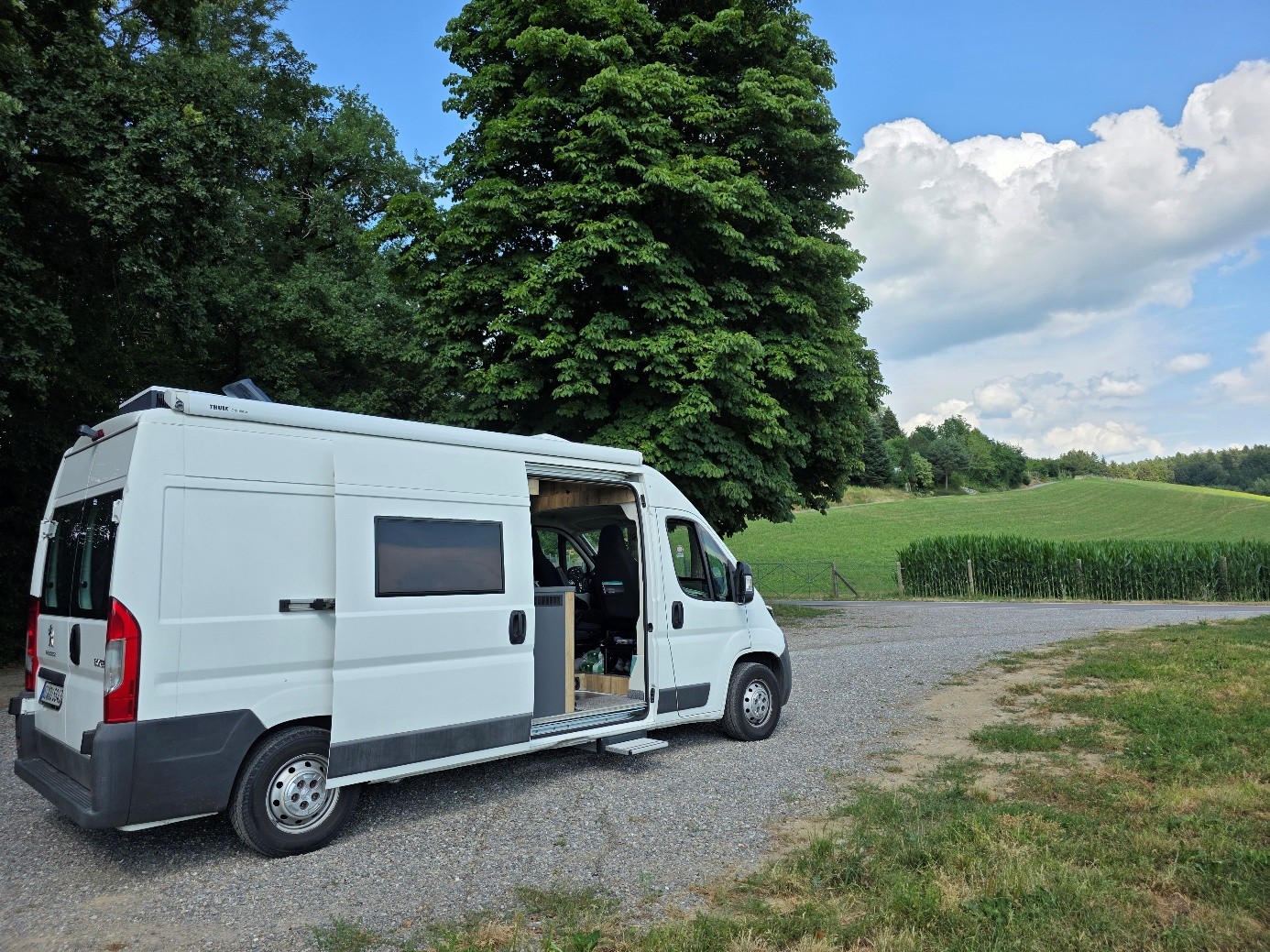 Weißer Camper mit ausgefahrener Trittstufe steht auf einem Feldweg zwischen Bäumen und Feldern.