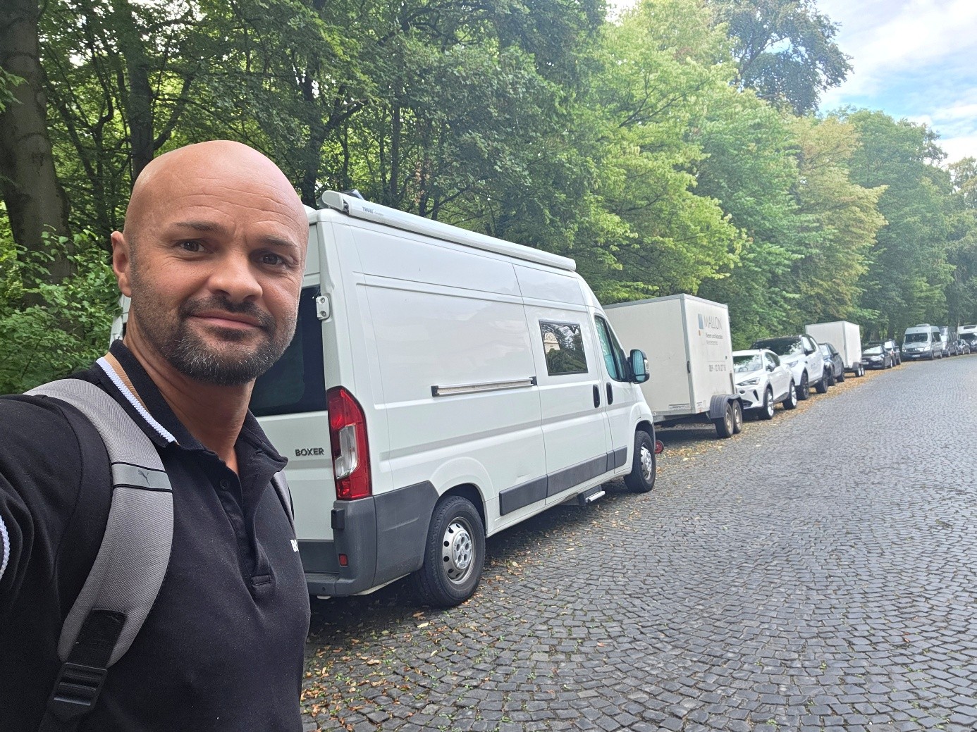 Eduard macht ein Selfie mit Rucksack vor seinem Camper in einer von Bäumen gesäumten Straße.