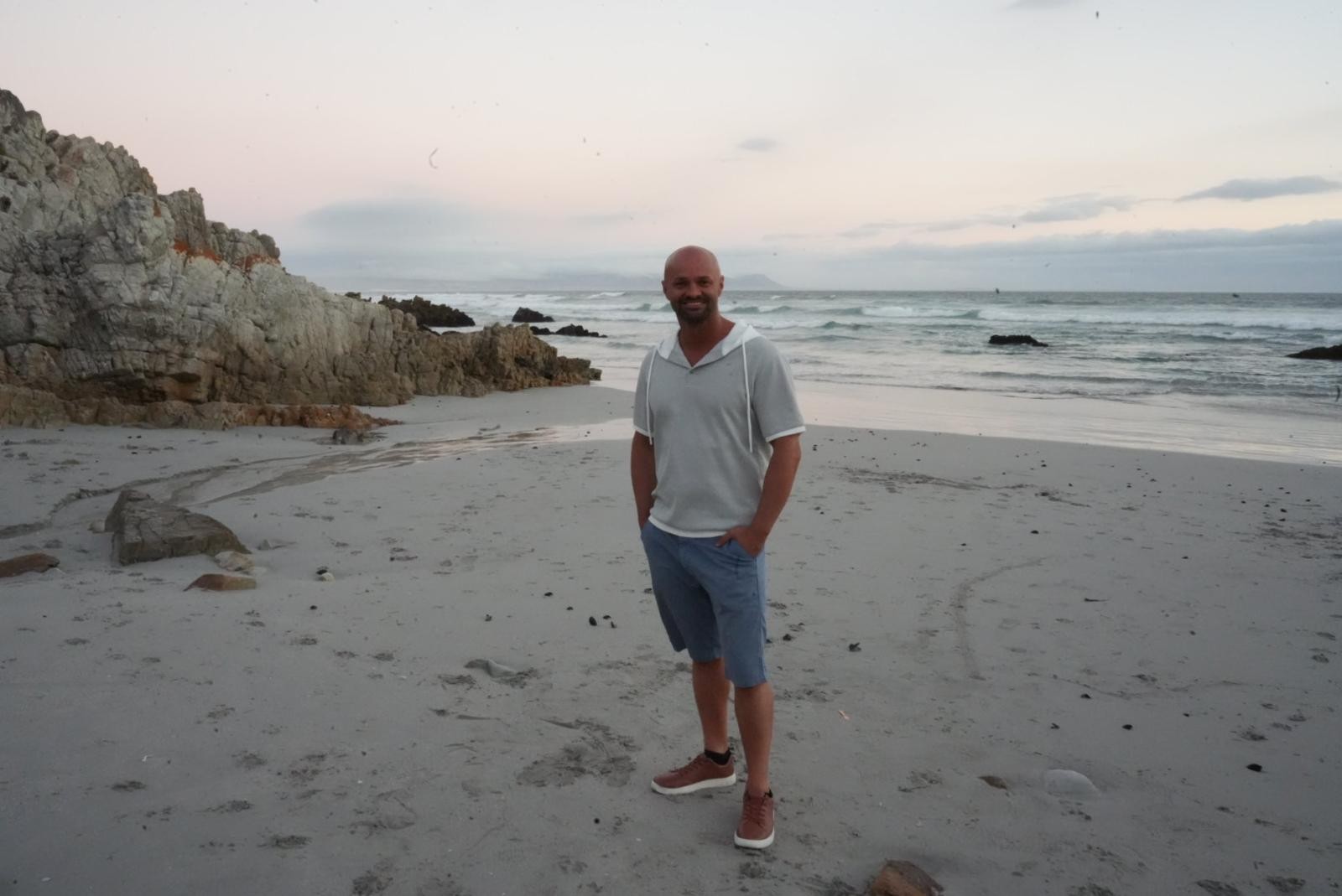 Ein Mann steht an einem Sandstrand mit Felsen und dem Ozean im Hintergrund.