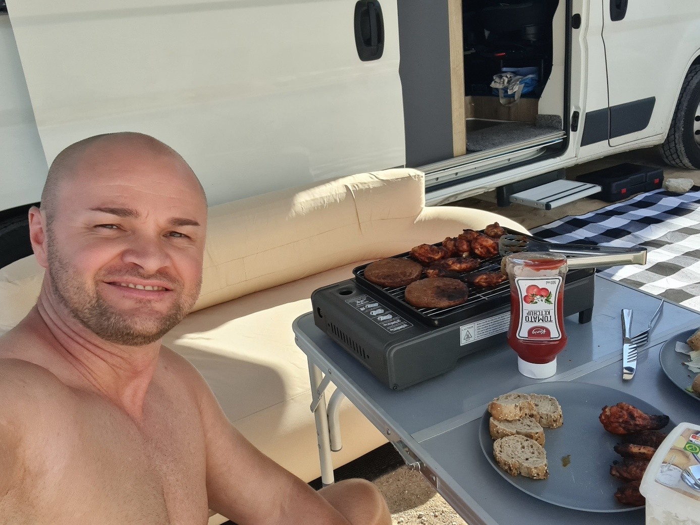 Eduard macht ein Selfie im Camper, vor ihm ein Tischgrill mit Fleisch und Brot.